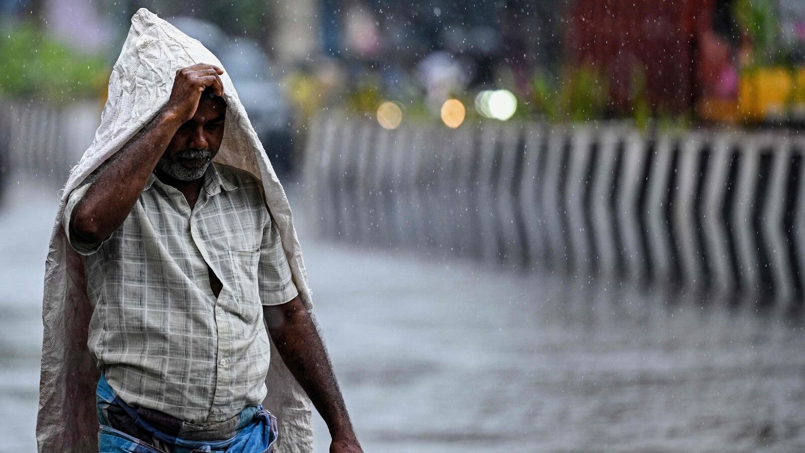 A man takes cover under a plastic sheet as he walks along a street during rainfall in Chennai on August 3, 2025.